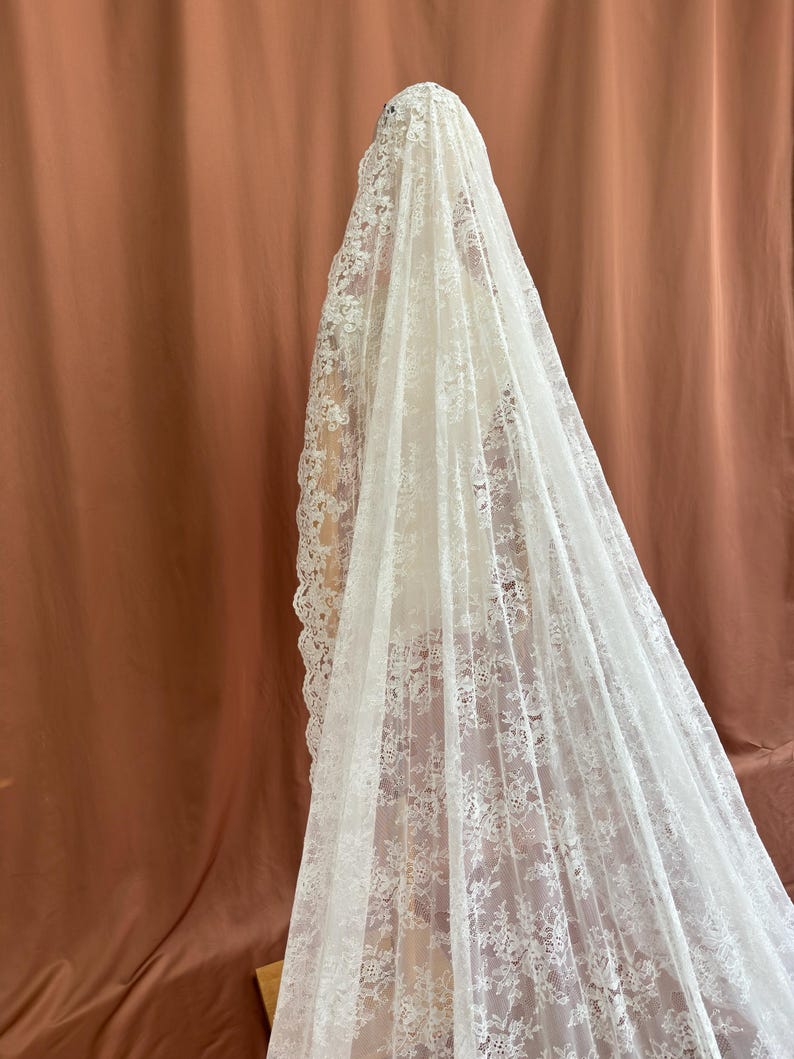 May include: A long, white lace veil with intricate floral patterns. The veil is draped over a mannequin, showcasing its delicate design and texture. The background is a solid, muted orange-brown.