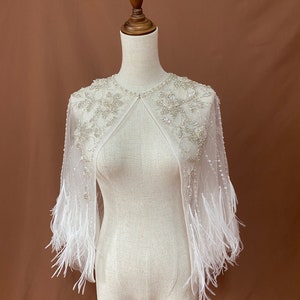 Puede incluir: Una capelina blanca y transparente con intrincados dise&ntilde;os florales con cuentas alrededor del cuello y los hombros. La capelina presenta mangas largas con adornos de plumas blancas en los pu&ntilde;os. La prenda se exhibe en un maniqu&iacute; sobre un fondo marr&oacute;n.