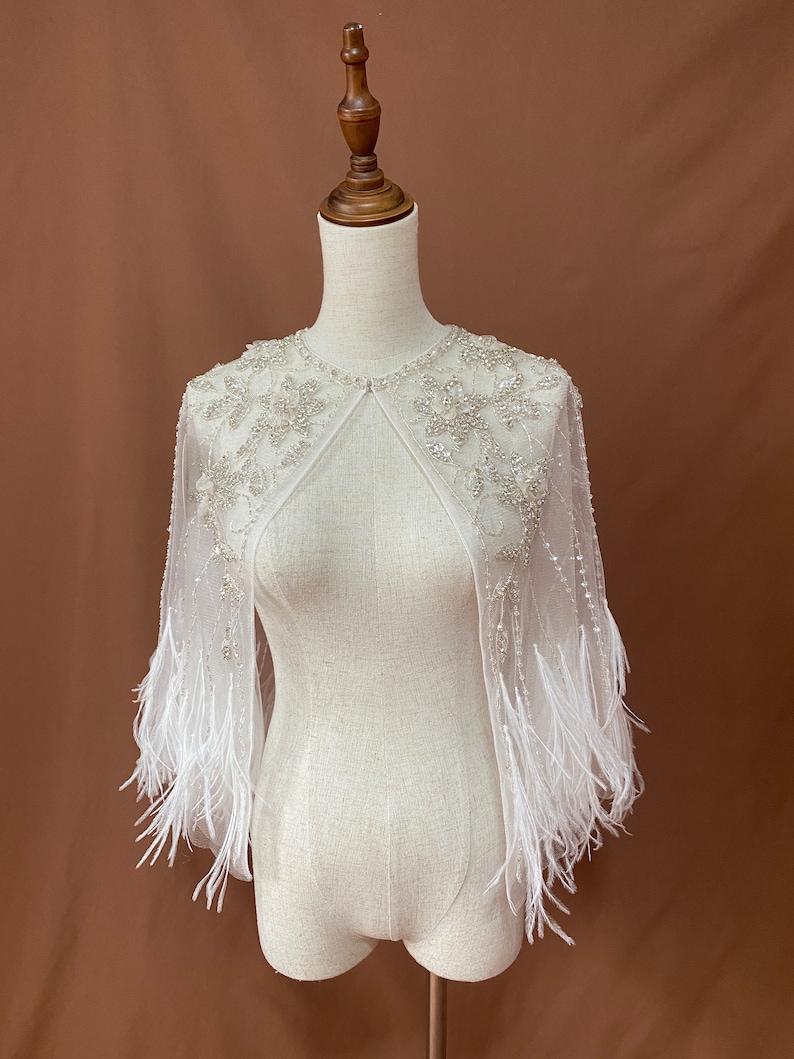 Puede incluir: Una capelina blanca y transparente con intrincados dise&ntilde;os florales con cuentas alrededor del cuello y los hombros. La capelina presenta mangas largas con adornos de plumas blancas en los pu&ntilde;os. La prenda se exhibe en un maniqu&iacute; sobre un fondo marr&oacute;n.