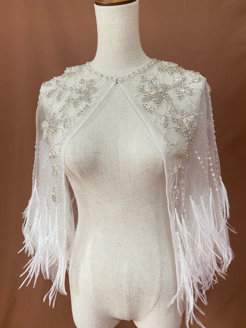 Puede incluir: Una capa blanca y transparente con intrincados adornos florales y de cuentas. La capa presenta delicados adornos de plumas a lo largo de las mangas, creando un aspecto elegante y et&eacute;reo. La capa se muestra en un maniqu&iacute;.