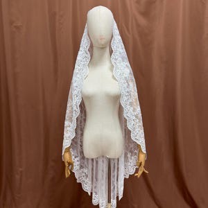 May include: A white lace veil with a scalloped edge is draped over a mannequin. The veil is made of delicate lace and has a floral pattern. The mannequin is on a wooden stand.