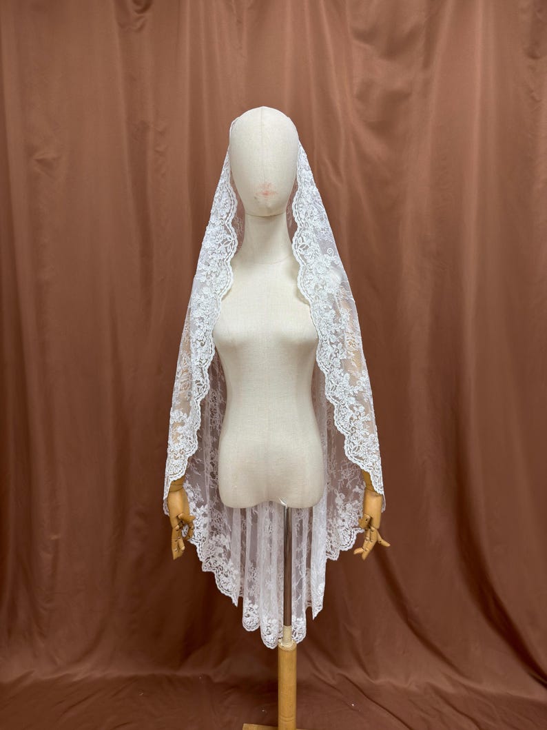 May include: A white lace veil with a scalloped edge is draped over a mannequin. The veil is made of delicate lace and has a floral pattern. The mannequin is on a wooden stand.
