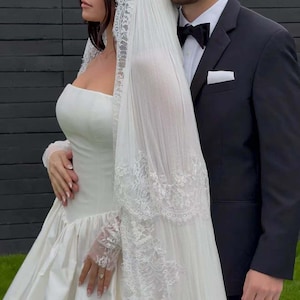 Puede incluir: Una novia con un vestido de novia de sat&eacute;n blanco con corpi&ntilde;o sin tirantes y falda amplia, combinado con un velo con ribete de encaje. Un novio con un esmoquin negro con pajarita est&aacute; a su lado.