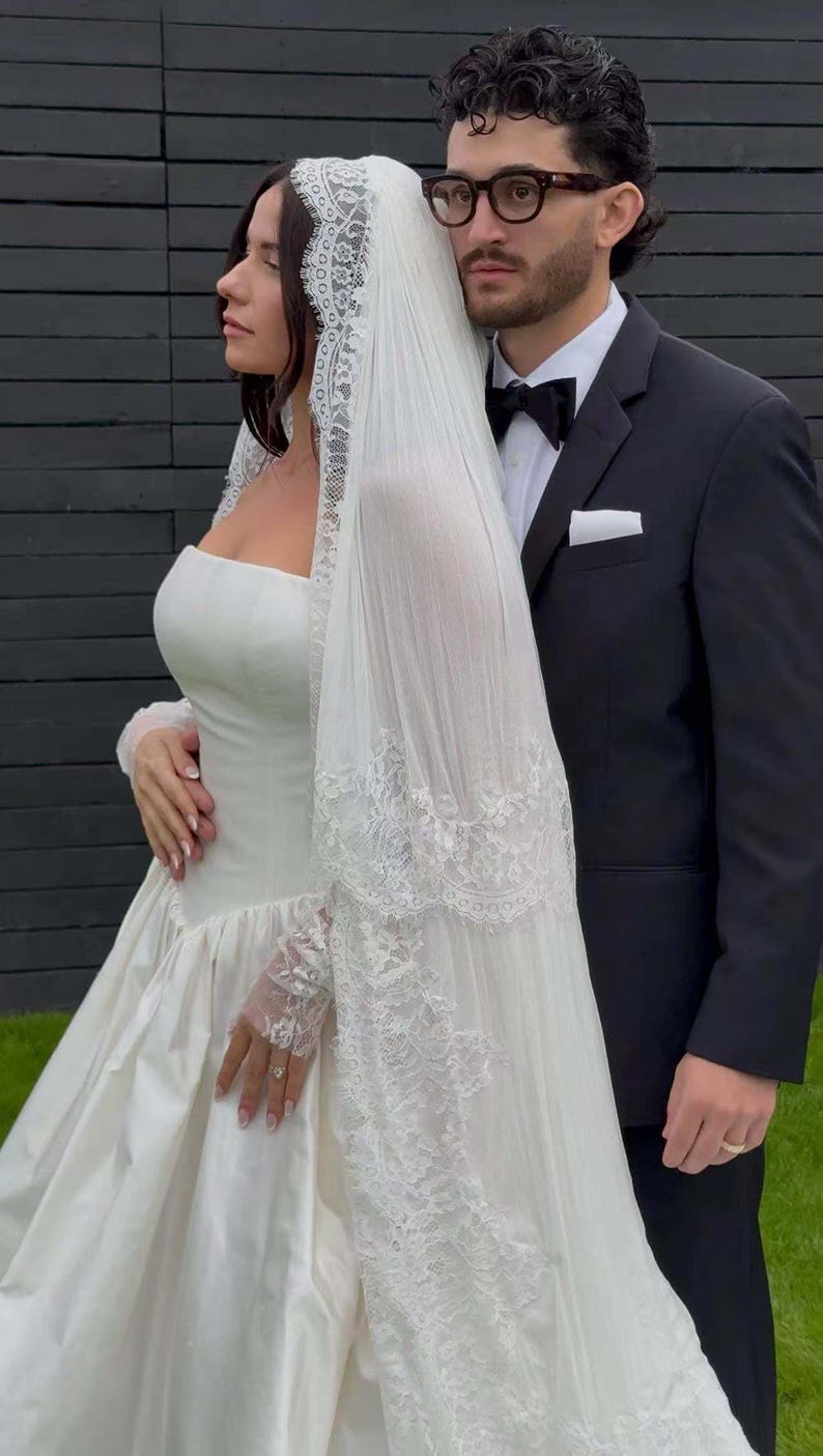 Puede incluir: Una novia con un vestido de novia de sat&eacute;n blanco con corpi&ntilde;o sin tirantes y falda amplia, combinado con un velo con ribete de encaje. Un novio con un esmoquin negro con pajarita est&aacute; a su lado.