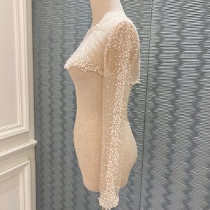 May include: A sheer, off-white bolero-style top with long sleeves, densely embellished with small, white beads. The garment is displayed on a mannequin, showcasing its delicate, textured design. The bolero is a formal wear item.