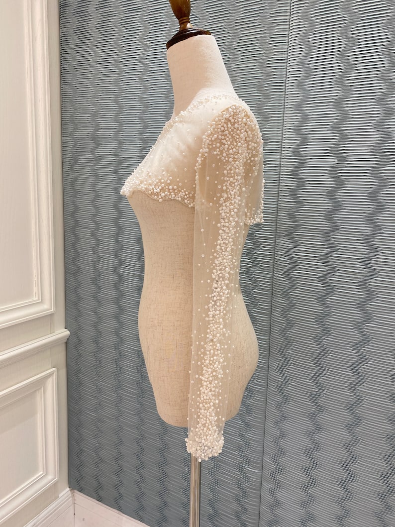 May include: A sheer, off-white bolero-style top with long sleeves, densely embellished with small, white beads. The garment is displayed on a mannequin, showcasing its delicate, textured design. The bolero is a formal wear item.