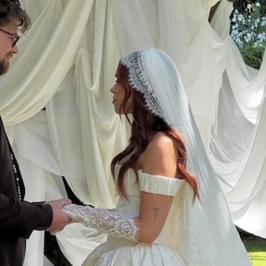 Puede incluir: Una novia con un vestido de novia blanco roto, con los hombros descubiertos, con una falda amplia y un velo largo. Lleva guantes de encaje y se da la mano con una persona con un traje negro. La ceremonia se lleva a cabo al aire libre, con tela blanca drapeada en el fondo.