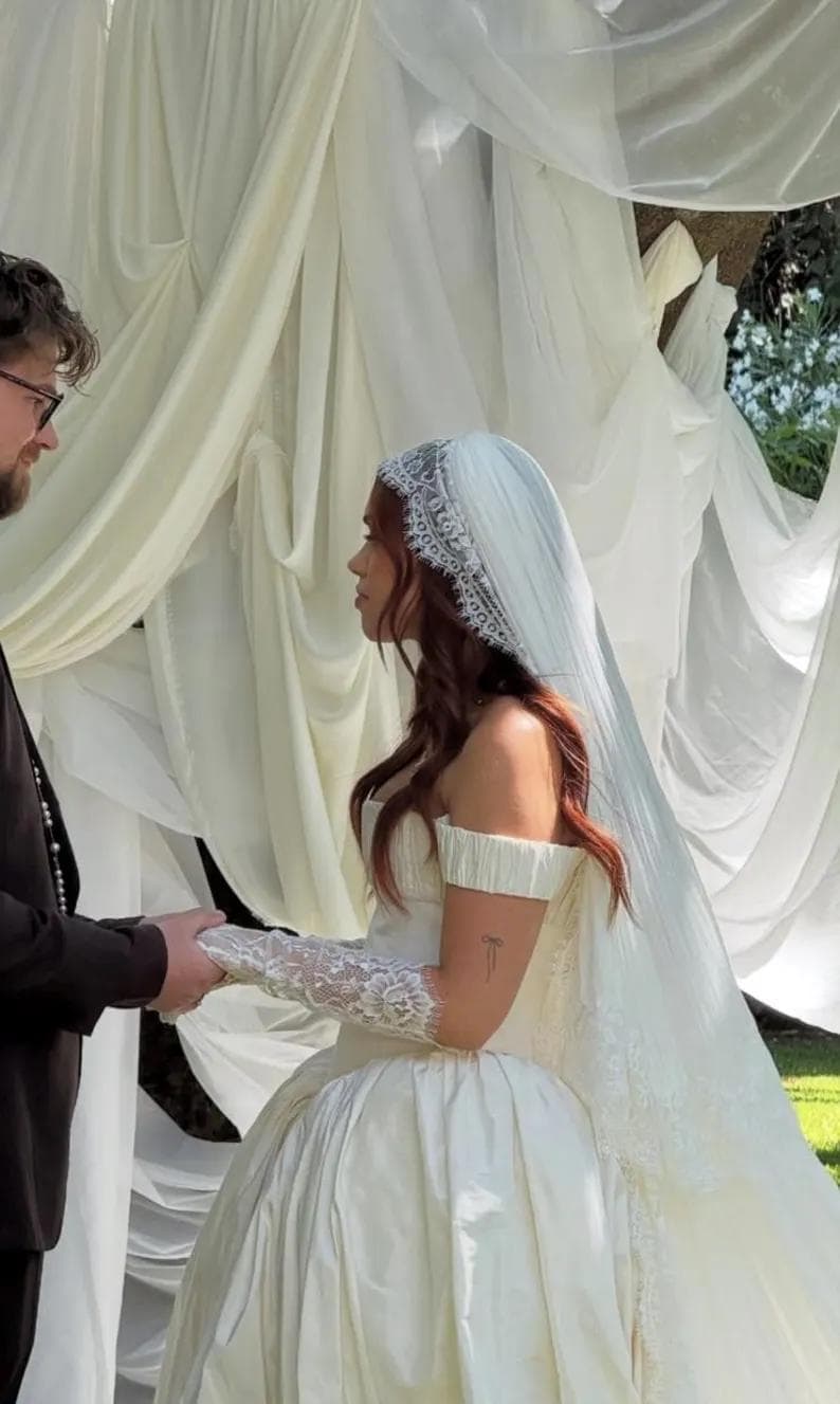 Puede incluir: Una novia con un vestido de novia blanco roto, con los hombros descubiertos, con una falda amplia y un velo largo. Lleva guantes de encaje y se da la mano con una persona con un traje negro. La ceremonia se lleva a cabo al aire libre, con tela blanca drapeada en el fondo.