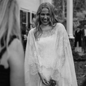 Puede incluir: Un vestido de novia blanco con un corpiño sin mangas y una capa de encaje transparente que llega hasta el suelo. La capa tiene mangas largas y detalles intrincados de encaje floral que caen sobre los hombros y los brazos, creando un aspecto superpuesto.