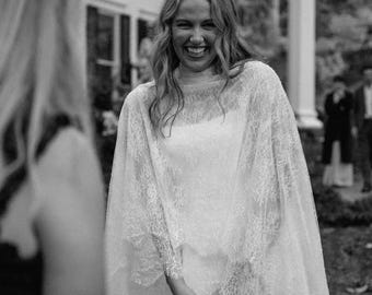 May include: A white wedding dress featuring a sleeveless bodice and a sheer, floor-length lace capelet overlay. The capelet has long sleeves and intricate floral lace detailing that drapes over the shoulders and arms, creating a layered appearance.