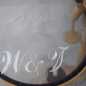 May include: A white embroidered fabric stretched across a wooden embroidery hoop. The text "zoeymade" is stitched at the top, with "W & T" in a cursive font below. The hoop has a dark blue trim.
