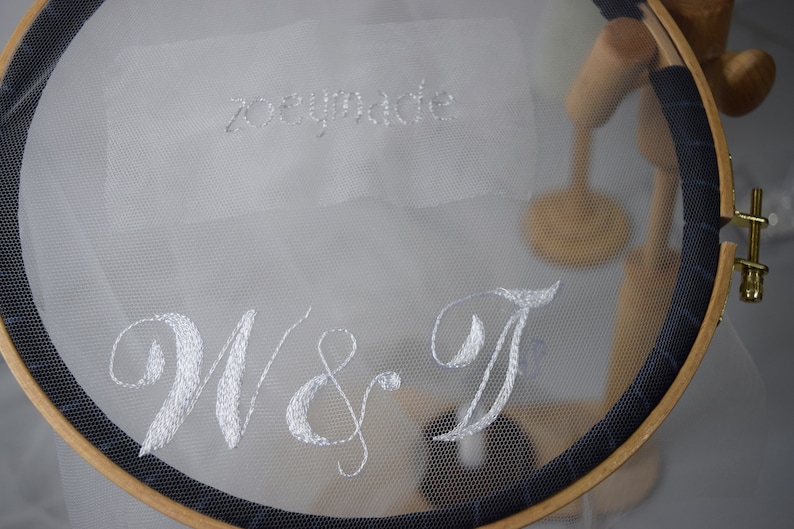May include: A white embroidered fabric stretched across a wooden embroidery hoop. The text "zoeymade" is stitched at the top, with "W & T" in a cursive font below. The hoop has a dark blue trim.