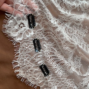 May include: White lace fabric with floral patterns and three black hair clips. The lace has a delicate, openwork design, and the clips are positioned across the fabric. The background is a solid brown color.
