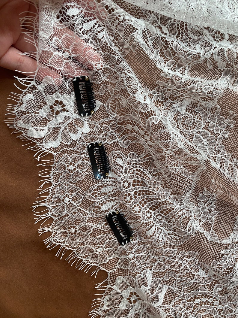 May include: White lace fabric with floral patterns and three black hair clips. The lace has a delicate, openwork design, and the clips are positioned across the fabric. The background is a solid brown color.