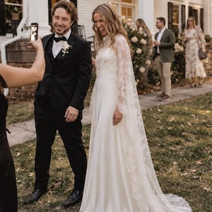 Puede incluir: Un vestido de novia blanco hasta el suelo con cuello alto y mangas largas de encaje transparente que se extienden en una cola. El vestido presenta detalles de encaje floral en el corpiño y las mangas, combinado con un esmoquin negro y pajarita.