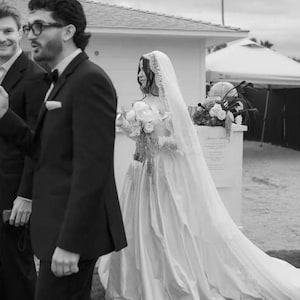 Puede incluir: Foto en blanco y negro de una boda. Una persona con un vestido de novia blanco con una larga cola y velo pasa junto a una persona con un traje negro. La novia lleva un ramo de flores.