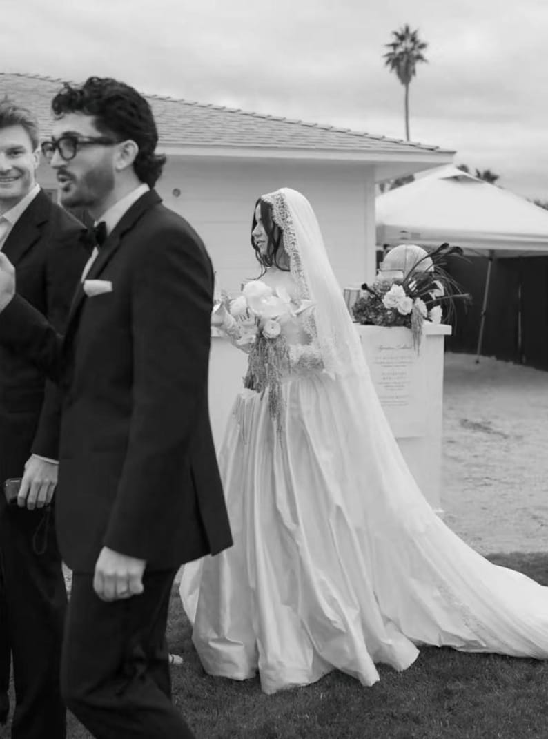 Puede incluir: Foto en blanco y negro de una boda. Una persona con un vestido de novia blanco con una larga cola y velo pasa junto a una persona con un traje negro. La novia lleva un ramo de flores.