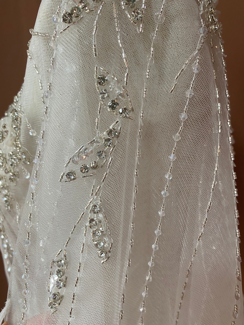 Puede incluir: Primer plano de una tela blanca y transparente adornada con cuentas plateadas y cristales transparentes. El dise&ntilde;o presenta l&iacute;neas de cuentas y grupos de cristales en forma de hojas, creando un patr&oacute;n delicado y brillante. La tela parece ser parte de un vestido de novia o de gala.