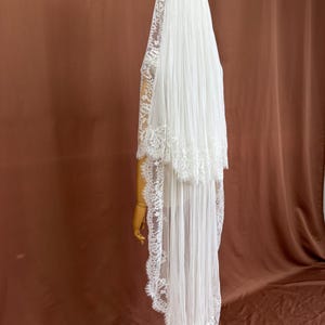Puede incluir: Un velo de novia blanco con un dise&ntilde;o en capas. El velo presenta un delicado ribete de encaje en los bordes y se exhibe en un maniqu&iacute; de madera. La tela parece transparente y fluida, creando un aspecto elegante.