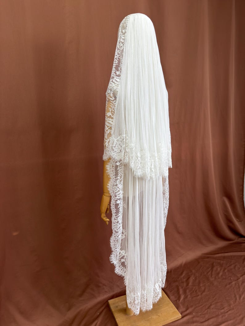 Puede incluir: Un velo de novia blanco con un dise&ntilde;o en capas. El velo presenta un delicado ribete de encaje en los bordes y se exhibe en un maniqu&iacute; de madera. La tela parece transparente y fluida, creando un aspecto elegante.
