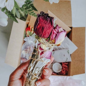 Könnte beinhalten: Ein in einer Hand gehaltenes Salbei-Smudge-Stick, verziert mit getrockneten Rosenblättern und -knospen. Der Stick ist mit Bindfaden umwickelt. Es ist Teil einer Geschenkbox mit einer Kerze und einem Beutel mit getrockneten Blumen. Die Box ist braun.