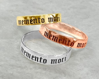 May include: Three stacking rings with the inscription "memento mori" in a gothic font. The rings are made of gold, rose gold, and silver.