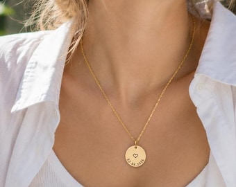 May include: A gold-toned necklace featuring a circular pendant with a small engraved heart symbol and the date 12.05.1975. The pendant hangs from a delicate gold-toned chain, resting against the chest area of a white ribbed tank top.