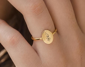 May include: A gold-toned ring featuring a small, oval-shaped flat plate. The center of the oval plate is engraved with a delicate, thin-lined botanical illustration of a single flower with a stem and leaves, centered on the metal surface.