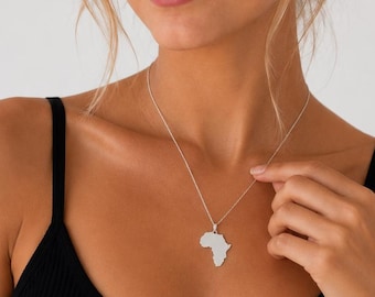May include: A silver-toned necklace featuring a pendant shaped like the continent of Africa. The pendant hangs from a delicate, thin silver chain. The piece is worn against a black ribbed tank top, highlighting the metallic finish of the jewelry.