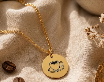 May include: A gold-toned necklace featuring a circular pendant with a black engraved graphic of a coffee cup on a saucer. The pendant is attached to a delicate gold-toned chain, creating a simple and elegant piece of jewelry for daily wear.