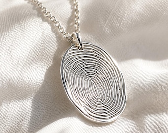 May include: A silver oval pendant necklace featuring a detailed, engraved fingerprint pattern on the front surface. The pendant is suspended from a delicate silver chain, showcasing a polished metallic finish with deep, etched lines.