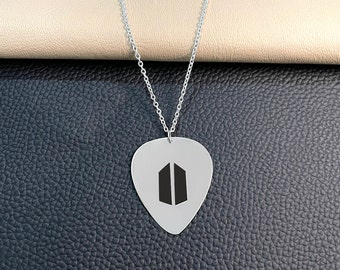 May include: Silver-toned guitar pick pendant necklace. The pendant features a black graphic design on a silver background. The necklace chain is also silver. The pendant is displayed on a textured black surface.