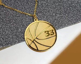 May include: A gold-colored basketball pendant necklace. The pendant features a basketball design with the number 33. The necklace chain is also gold-colored. The pendant is round and has a polished finish.
