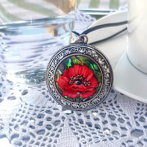 May include: A silver pendant necklace featuring a vibrant red poppy flower design. The pendant has an ornate, detailed border and a black cord. The necklace rests on a white lace doily, with a glass of water and a cup of coffee in the background.
