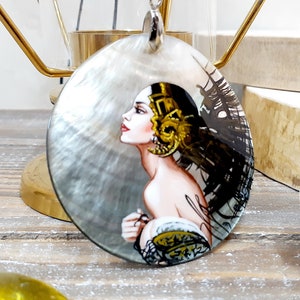 May include: A pendant necklace featuring a hand-painted portrait of a woman on a large, oval-shaped shell. The woman has dark hair and is adorned with gold headwear. The shell has a pearlescent sheen, and the pendant hangs from a black cord.