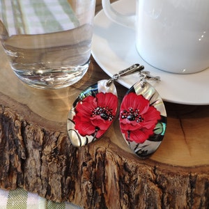 May include: Oval-shaped earrings with a red poppy design. The earrings have a silver-colored hook and are displayed on a wooden surface. A cup of coffee and a glass of water are in the background.