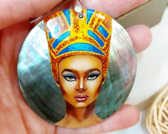 May include: A round, iridescent, mother-of-pearl pendant with a painted portrait of a woman wearing a gold and blue crown. The pendant is on a black cord.