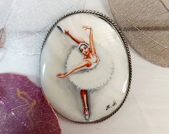 May include: Oval-shaped brooch featuring a hand-painted ballerina in a white tutu, poised mid-leap. The brooch has a silver-toned, rope-like border. The ballerina is depicted with red ballet shoes and a delicate expression.
