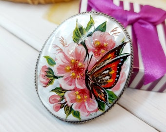 May include: A silver oval brooch featuring a painted design of pink flowers and a butterfly with orange, brown and black wings.
