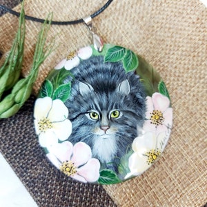 May include: A round pendant necklace with a painted image of a gray cat with green eyes surrounded by white and pink flowers. The pendant is on a black cord.