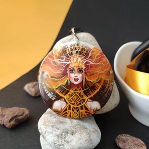 Könnte beinhalten: Runder Anhänger mit einem gemalten Porträt einer Frau mit orangefarbenem Haar und einem goldenen Kopfschmuck. Der Anhänger hat eine silberne Öse und eine schwarze Kordel. Die Frau hat blaue Augen und rote Lippen. Der Anhänger liegt auf einem weißen Stein.