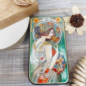 May include: A vintage-style tin box with a colorful Art Nouveau design featuring a woman with flowing hair and a floral motif. The box has a green background with gold accents and the artist's signature "Mucha" at the bottom.