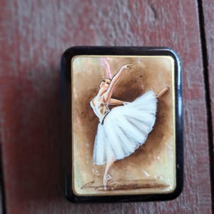 May include: A small, rectangular, black box with a painted image of a ballerina in a white tutu. The ballerina is in a graceful pose, with one arm raised. The background is a warm, neutral tone.