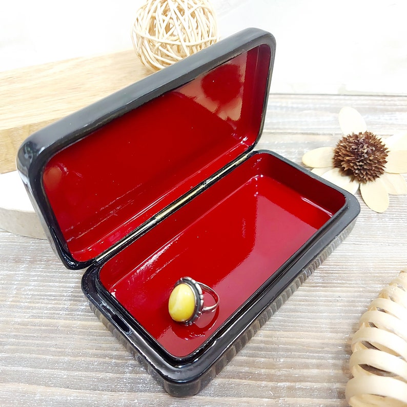 May include: A black lacquered wooden box with a red velvet interior. The box is open and a yellow oval ring is inside.