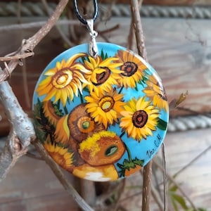 May include: A round pendant with a hand-painted design of sunflowers in shades of yellow and brown against a light blue background. The pendant hangs from a black cord. The artist's signature is visible.