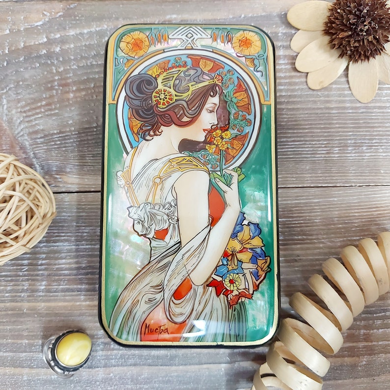 May include: A rectangular wooden box with a hinged lid. The lid features a colorful Art Nouveau style illustration of a woman with flowing hair and a floral design. The box is signed "Mucha" in the lower right corner.