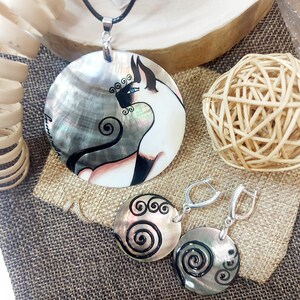 May include: A pendant necklace and matching earrings set. The pendant is a large, round, iridescent shell with a hand-painted cat design. The earrings are round shells with a spiral design. The jewelry is displayed on a burlap surface.