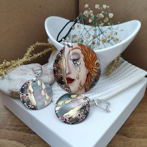 Peut inclure: Ensemble de bijoux faits à la main comprenant un pendentif et des boucles d'oreilles. Le pendentif représente un visage de femme peint avec des larmes, des lèvres rouges et des accents dorés. Les boucles d'oreilles et le pendentif sont ronds avec des détails dorés et rouges.