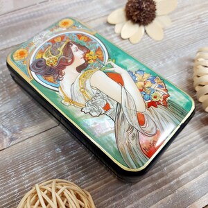 May include: A rectangular wooden box with a green background and a painted image of a woman in a white dress holding flowers. The box is decorated with gold trim and the artist's signature, "Mucha", is visible in the lower right corner.