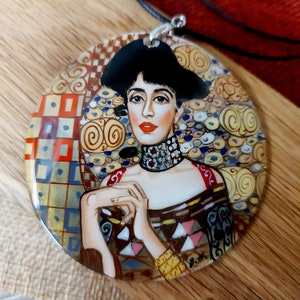 May include: Round pendant featuring a portrait of a woman in the style of Gustav Klimt. The pendant has a black cord and silver-tone bail. The woman has red lips, a black hat, and a patterned dress with gold, pink, and blue accents. The background has gold and blue swirls.